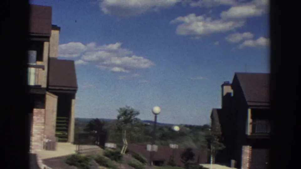 1972 Toronto home-movie frame showing the edge of a residential complex with landscaped grounds, trees, round lamps, and a bright clouded sky.