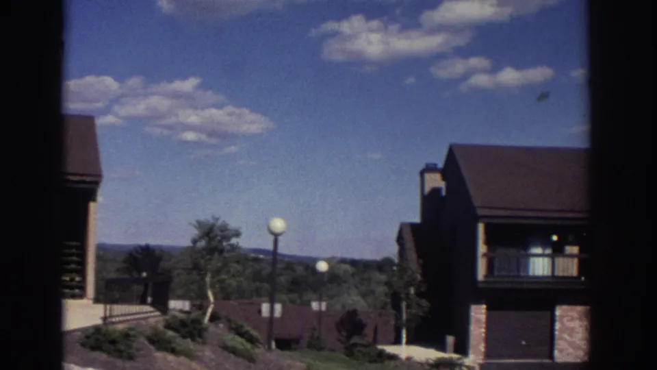 1972 Toronto home-movie frame showing low-rise townhouse-style buildings, globe lamps, landscaping, and distant tree-covered land.