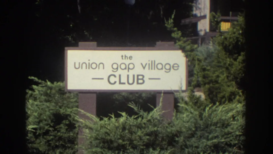 1972 Toronto frame centered on a sign reading the Union Gap Village Club, surrounded by dense landscaping and partial buildings.