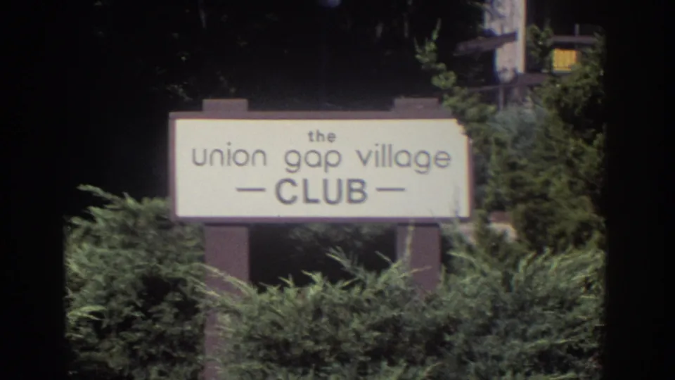 Home-movie frame from 1972 Toronto showing the Union Gap Village Club sign partly framed by shrubs and dark film edges.