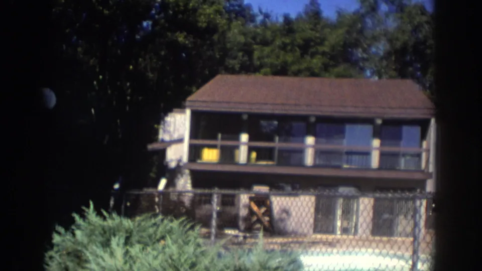 1972 Toronto exterior showing a low-rise residential building, balcony railing, chain-link fence, shrubs, and possible pool-area equipment.
