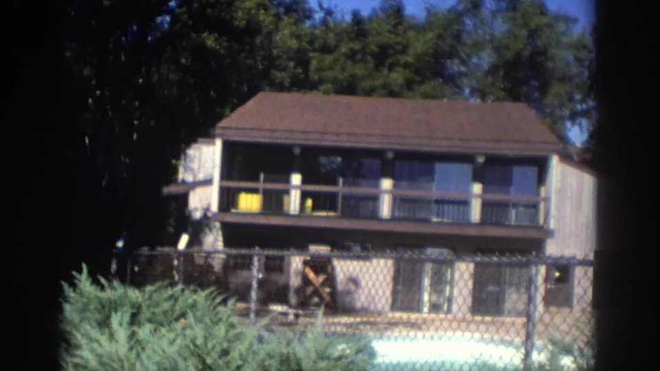 1972 Toronto 8mm frame of a two-story house with balcony, large windows, chain-link fence, shrubs, and a pool in front.
