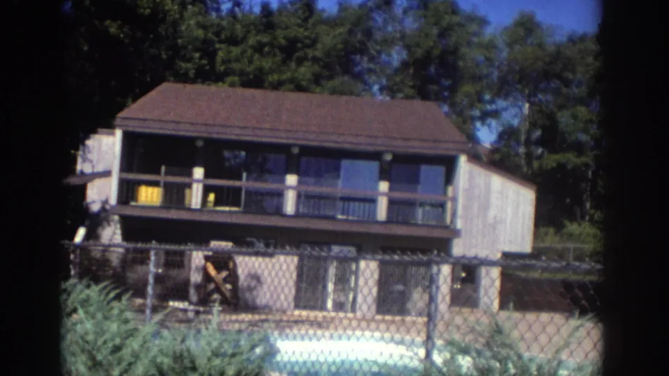 1972 Toronto home-movie frame showing the house facade, upper balcony, chain-link fence, pool edge, and trees in bright daylight.