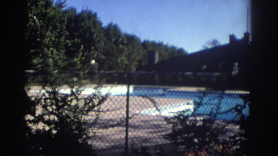 1972 Toronto home-movie frame focused on the pool behind a chain-link fence, with trees, shrubs, blue sky, and a shaded structure at right.