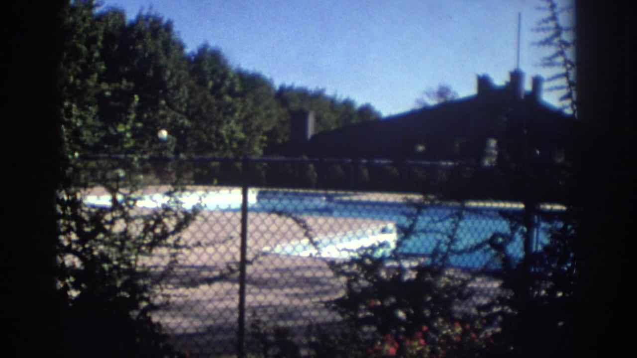 1972 Toronto home-movie frame showing a chain-link fence, bright outdoor pool, trees, and a dark building in the background.