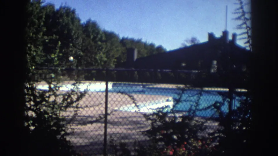1972 Toronto home-movie frame showing a chain-link fence, bright outdoor pool, trees, and a dark building in the background.