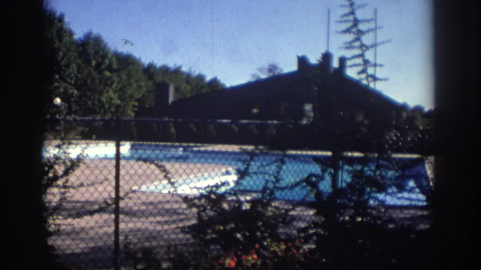 Fenced pool area in Toronto, 1972, with saturated blue water, surrounding trees, and a dark roofline in the background.