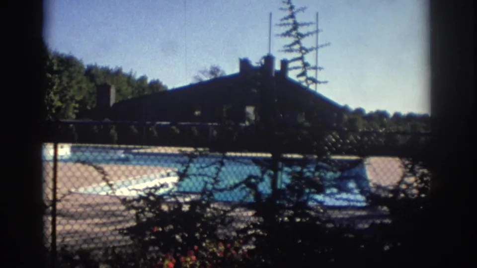 Soft 8mm view of a fenced outdoor pool in Toronto, 1972, with trees, deck pavement, and a shadowed building behind it.