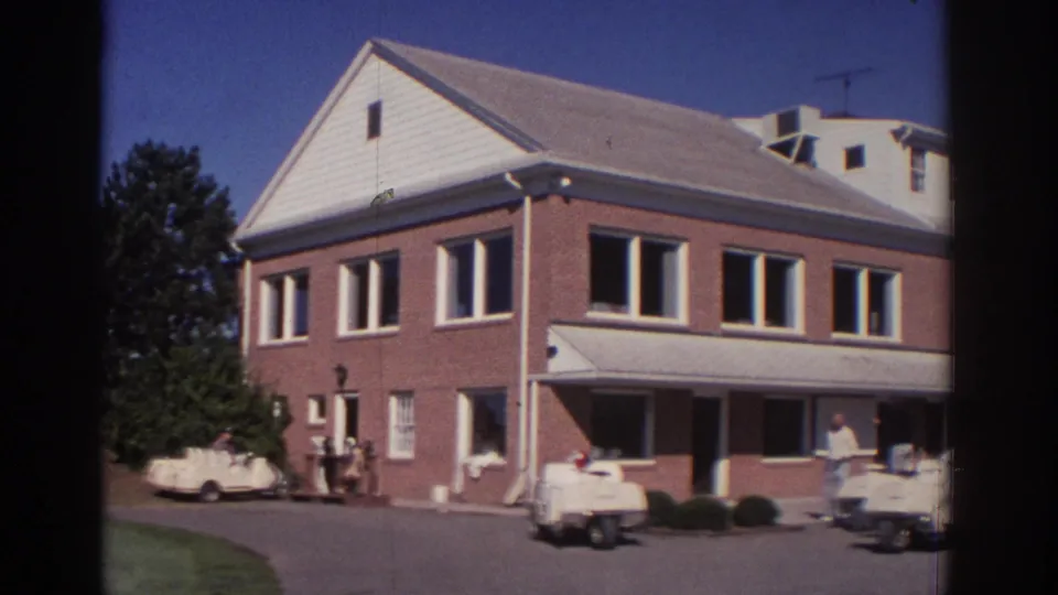 1972 Toronto exterior of a brick building with a white gabled roof, covered awning, golf carts, and people near the entrance.