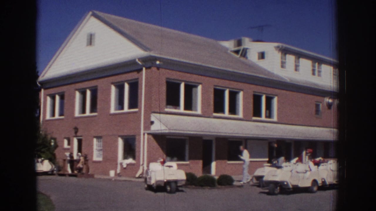 1972 Toronto home-movie frame showing a brick golf-course building with a white awning and golf carts lined up on the paved forecourt.