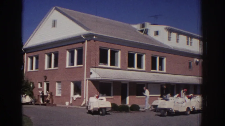 1972 Toronto home-movie frame showing a brick golf-course building with a white awning and golf carts lined up on the paved forecourt.