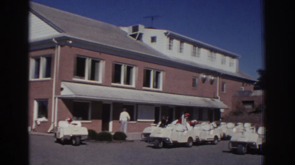 1972 Toronto footage frame with white golf carts in rows outside a brick building, with an adult visible near the entrance area.