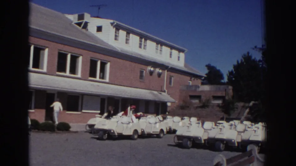 1972 Toronto home-movie frame showing a closer angle on a brick golf facility with several white carts parked along the paved area.