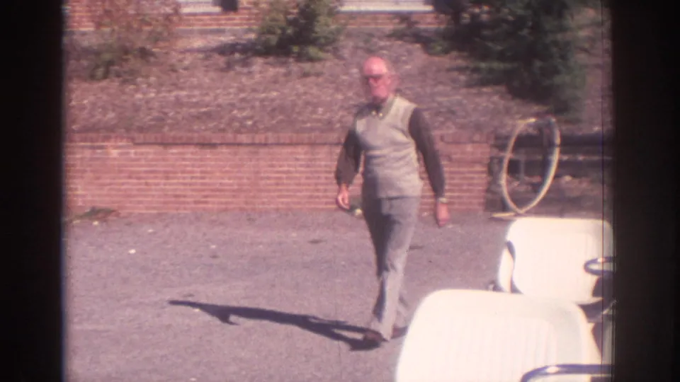 Adult man wearing glasses, a light vest, dark shirt, and trousers walks on a paved area near brickwork and part of a golf cart.
