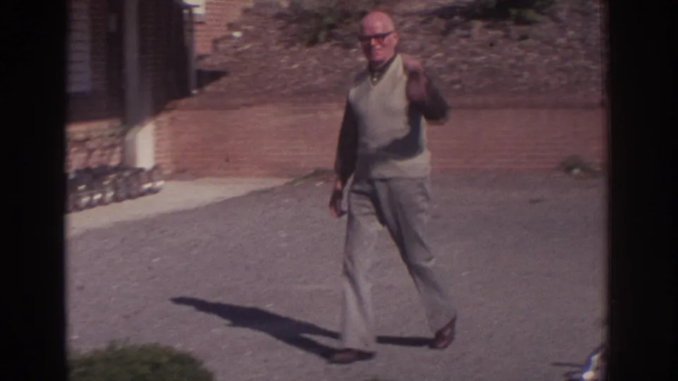Adult man in glasses and a light vest raises one hand while standing on a paved drive near a brick and stone building exterior.