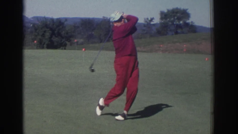 1972 Toronto home-movie frame showing a man in a red tracksuit completing a golf swing with trees and hills behind him.