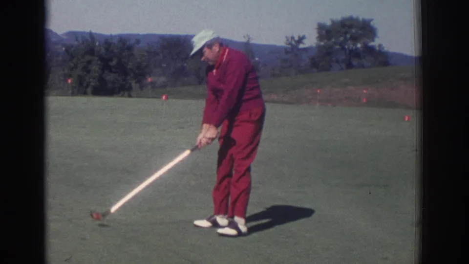 1972 Toronto home-movie frame of a red-clad golfer near impact or setup, with tee markers, trees, and hills in the background.