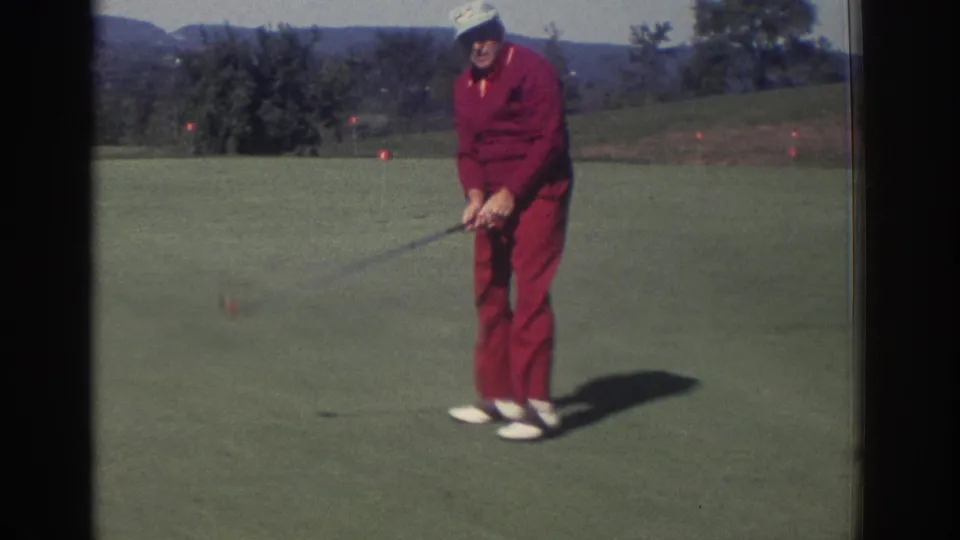 1972 Toronto 8mm frame of an adult man in a red tracksuit and cap addressing a golf ball on a green.