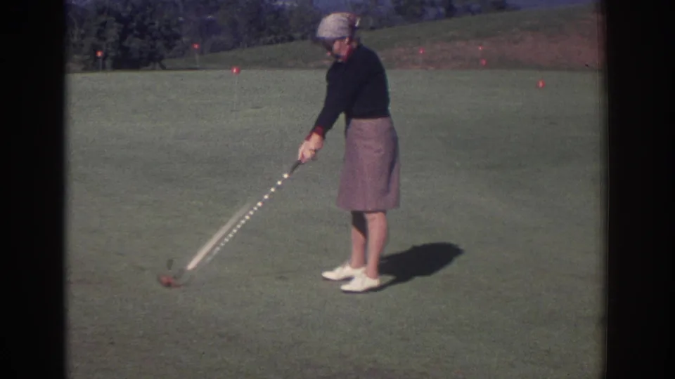1972 Toronto 8mm frame of an adult woman in a dark sweater, skirt, and headscarf swinging a golf club.