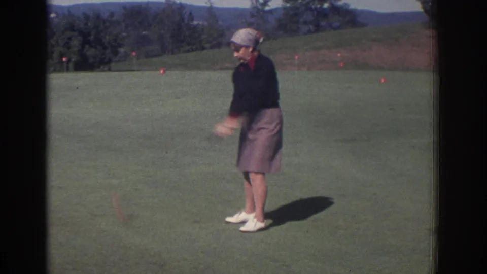 1972 Toronto 8mm frame showing an adult woman in skirt and headscarf after a golf swing on the course.
