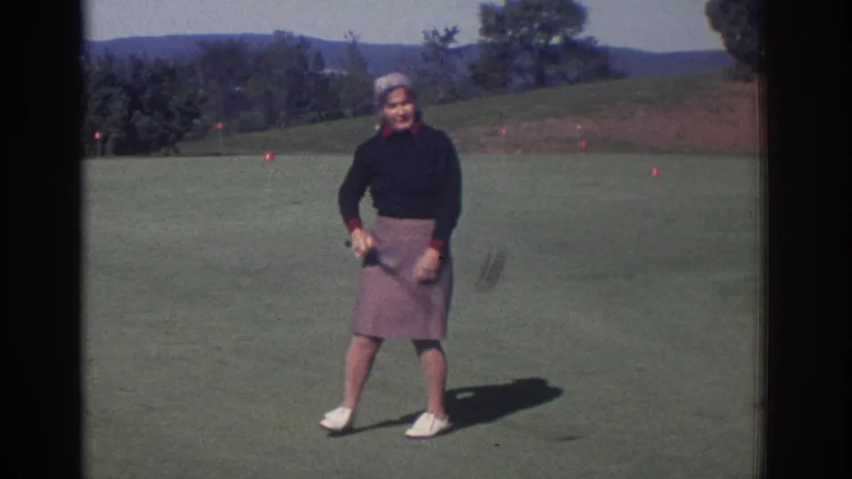 1972 Toronto 8mm frame of an adult woman smiling toward the camera while holding a golf club on the course.