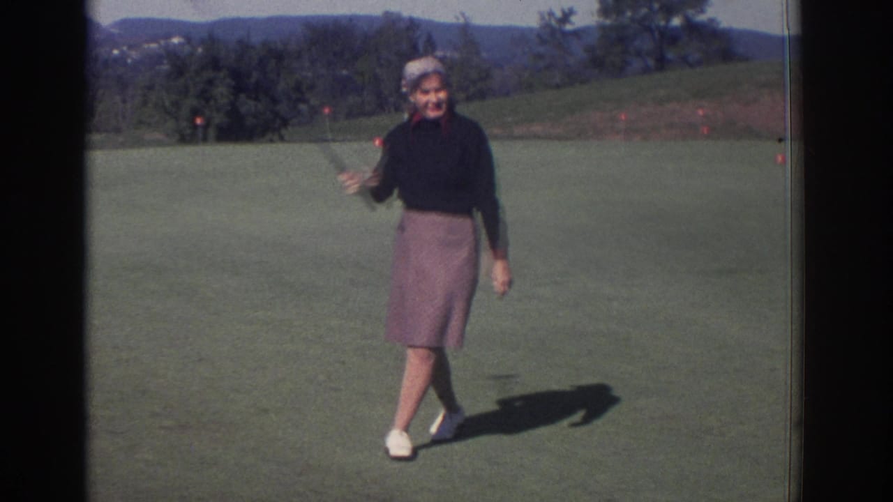 Woman holding a golf club on a sunlit green with trees and hills behind her.