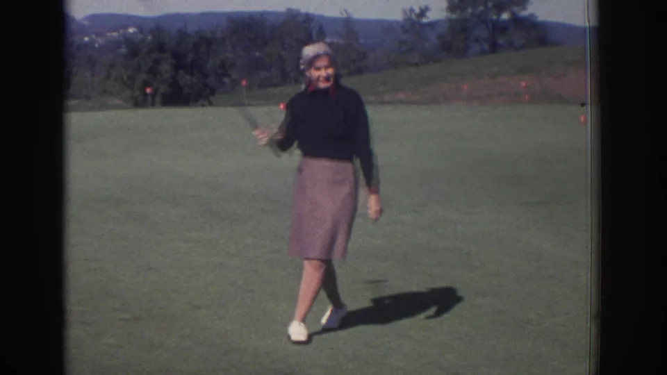 1972 Toronto home-movie frame showing a smiling woman with a golf club on a sunny golf course, with trees and distant hills behind her.