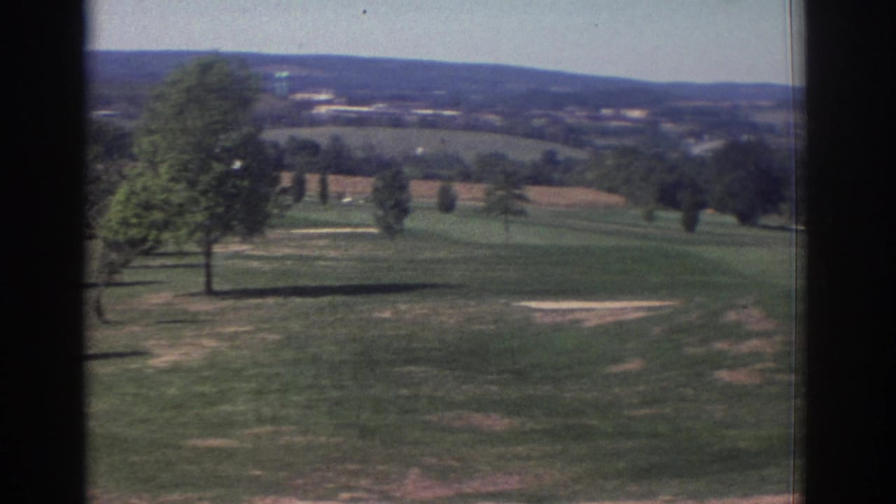 Open golf course fairway with trees, bare patches, and distant hills under clear sky.