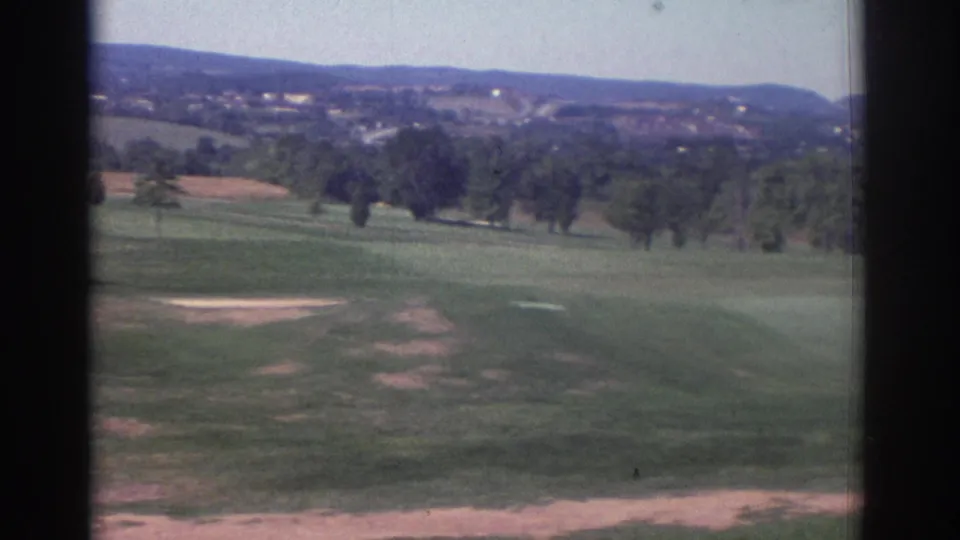 1972 Toronto home-movie frame showing a broad golf course view with green fairway, scattered trees, pale ground patches, and low hills beyond.