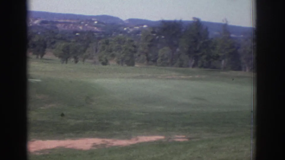 1972 Toronto home-movie frame of a quiet golf course landscape, with green turf, tree lines, pale ground patches, and hills in the background.