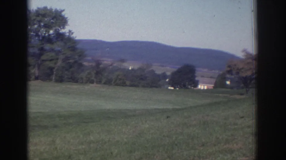 1972 home-movie frame of a golf course fairway with trees, distant low buildings, and hills beyond.