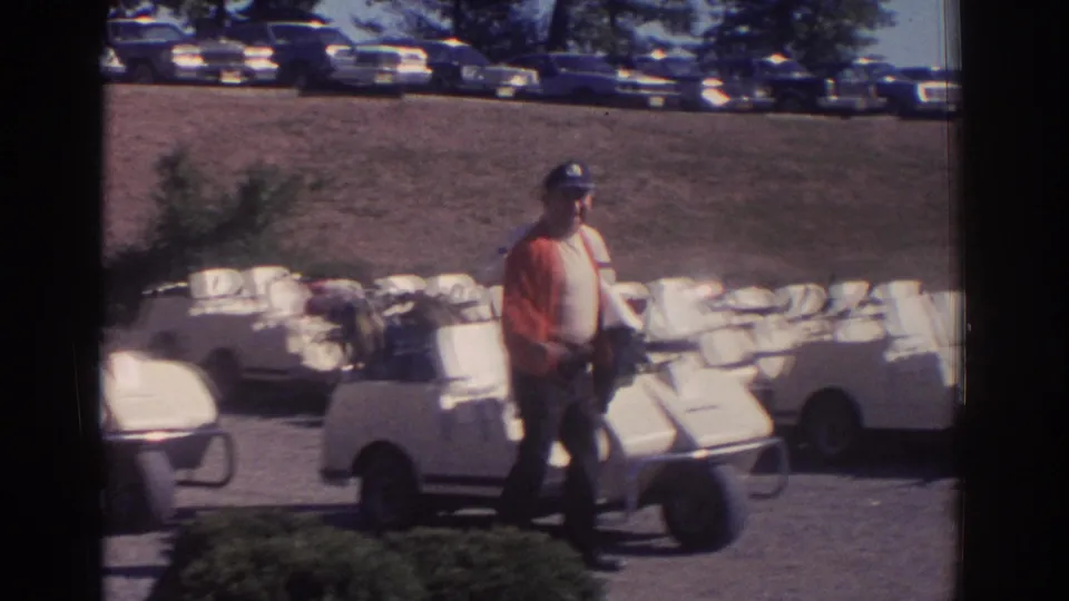 Adult man in a red jacket and blue cap stands beside a white golf cart at a 1972 Toronto golf course, with parked cars behind.