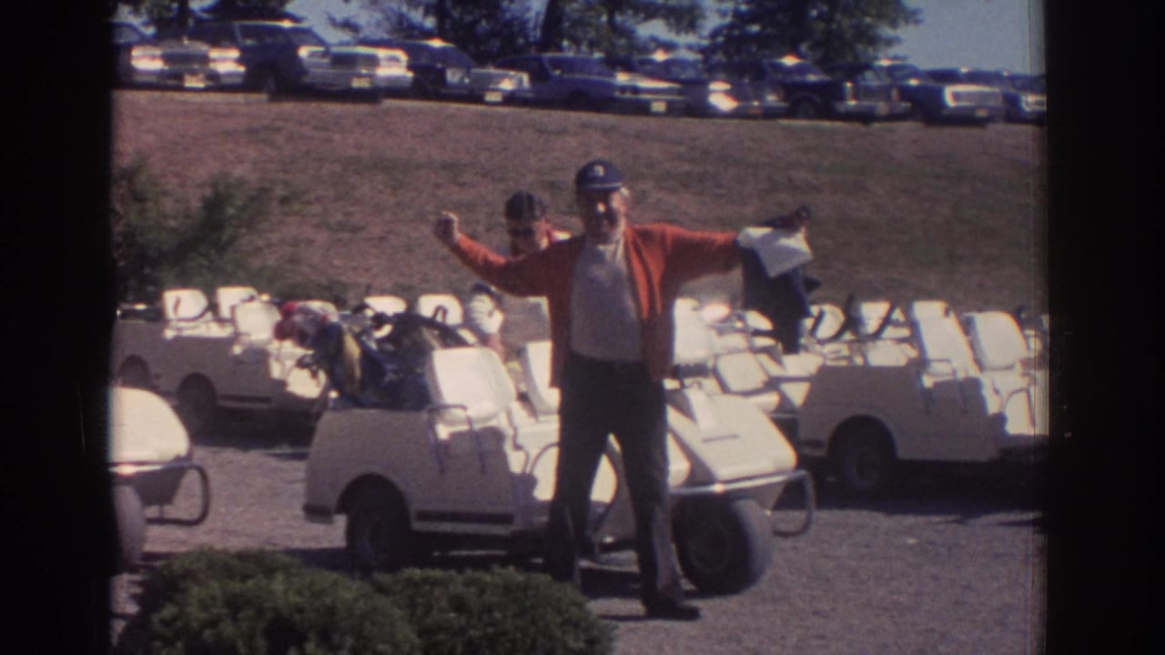 In supported 1972 Toronto golf club footage, a man in a red cardigan and cap gestures beside rows of white golf carts.