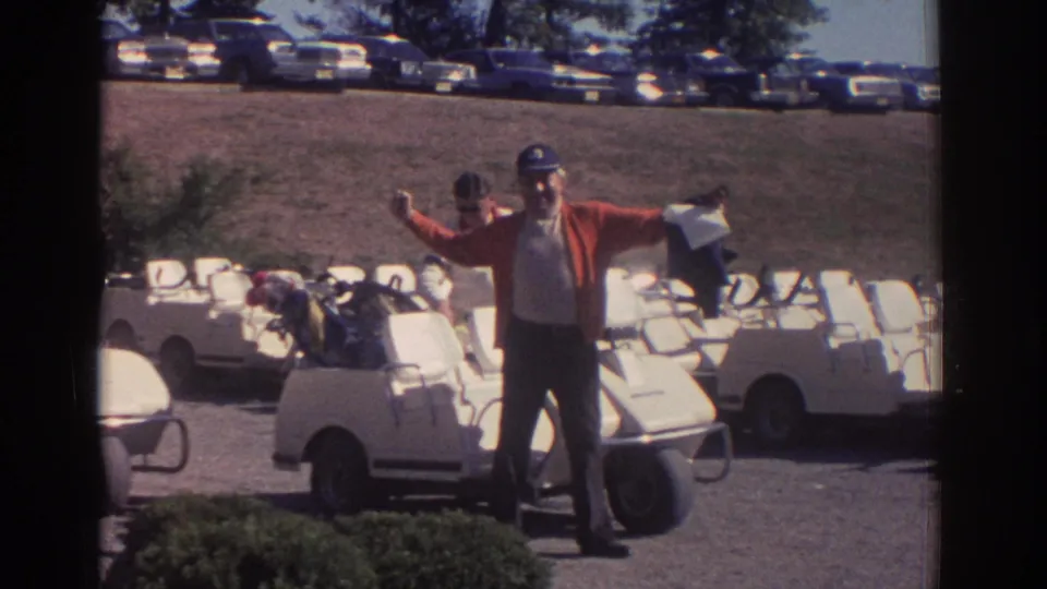 In supported 1972 Toronto golf club footage, a man in a red cardigan and cap gestures beside rows of white golf carts.