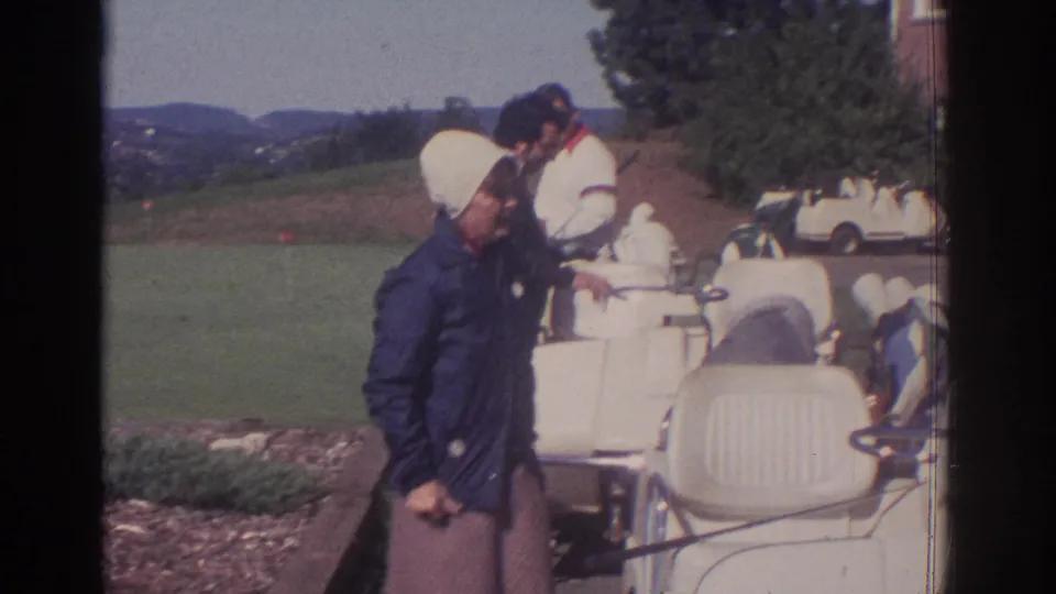 A woman in a white hat and blue jacket stands near white golf carts as another adult leans over a cart at the golf club.