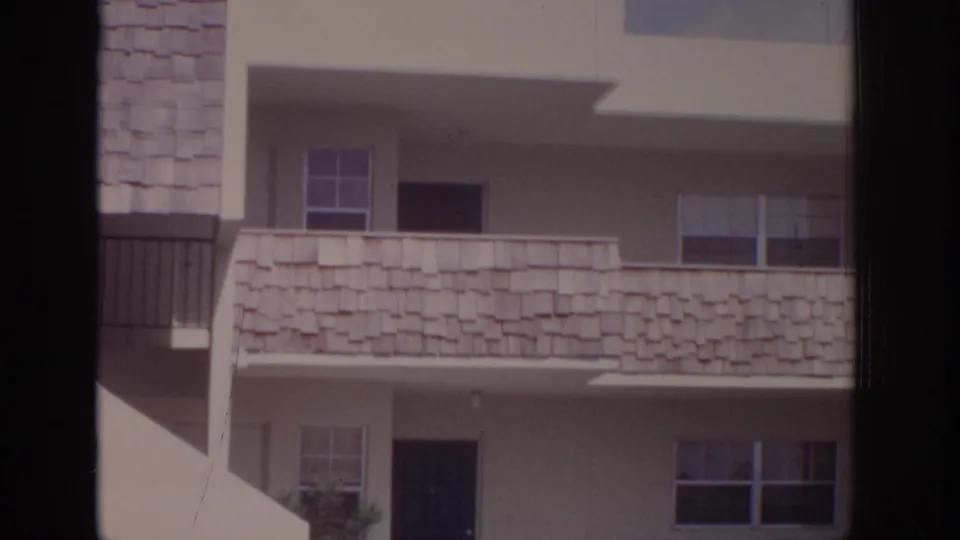 1973 Naples, Florida apartment exterior with white stucco walls, shingled balcony panels, dark doors, windows, and a small landscaped edge.
