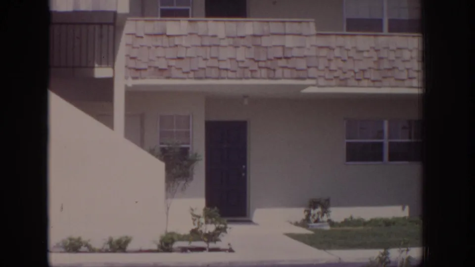 1973 Naples, Florida residential exterior showing white apartment units, a stair wall, dark door, windows, small lawn, and sparse landscaping.