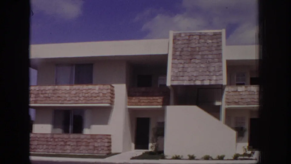 1973 Naples, Florida apartment exterior with white stucco, shingled balcony panels, a stair wall, dark doorway, windows, and sparse shrubs.