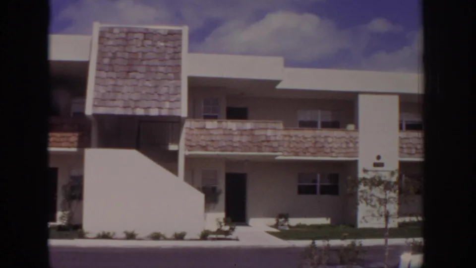 Naples, Florida, 1973 frame showing the apartment complex mid-pan, with white stucco, shingled balcony fronts, and a young tree near the walkway.