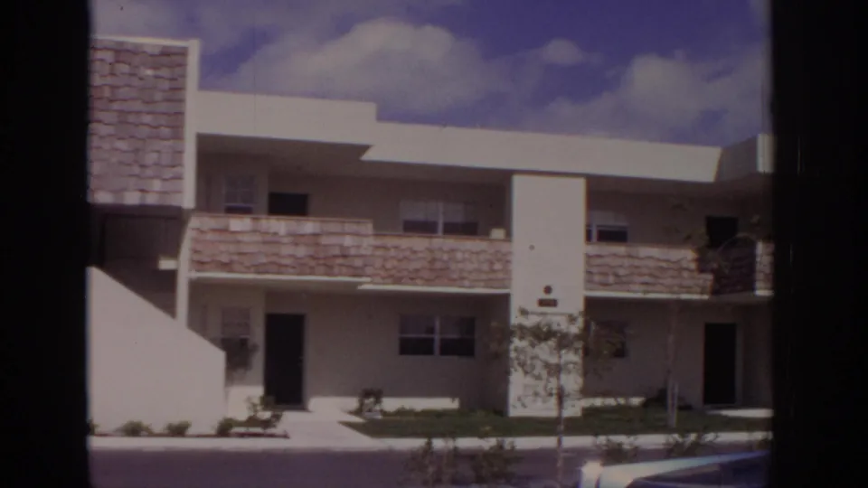 Final 1973 Naples frame of the apartment exterior, showing shingled balcony fronts, dark doorways, a young tree, and part of a car at the edge.