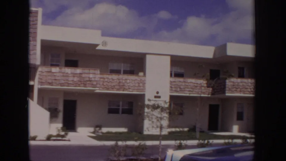 Naples, Florida, 1973 view of a white two-story residential building with flat roofs, stone-textured balcony panels, lawn, and parked car edge.