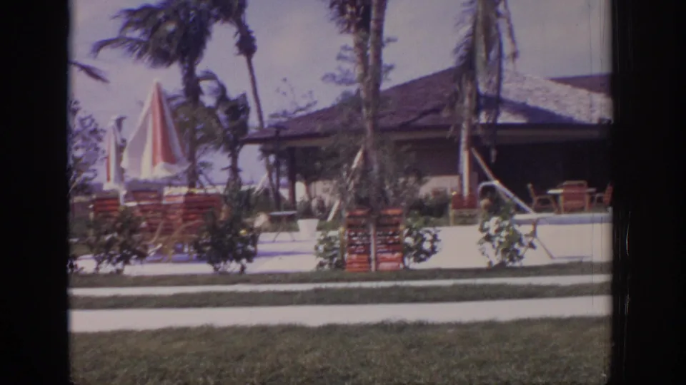 1973 Naples, Florida frame showing a patio area with red-and-white striped umbrellas, lounge chairs, palms, lawn, and a low building.