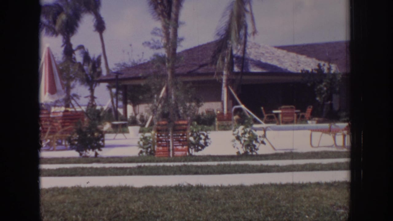 Poolside resort scene with lounge chairs, striped umbrella, palms, lawn, and low roofed building.