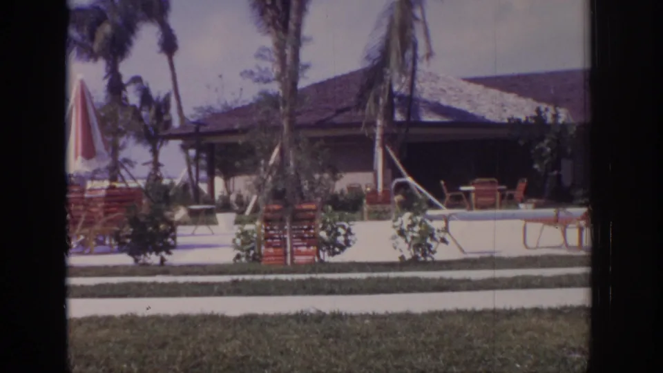 1973 Naples, Florida home-movie frame showing a resort pool area with red lounge chairs, a striped umbrella, palms, shrubs, and a low tiled-roof building.