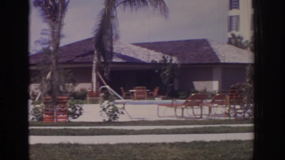 Naples, Florida resort poolside in 1973 with red chairs around the pool, palms and landscaping, a low roofed structure, and part of a taller hotel building.