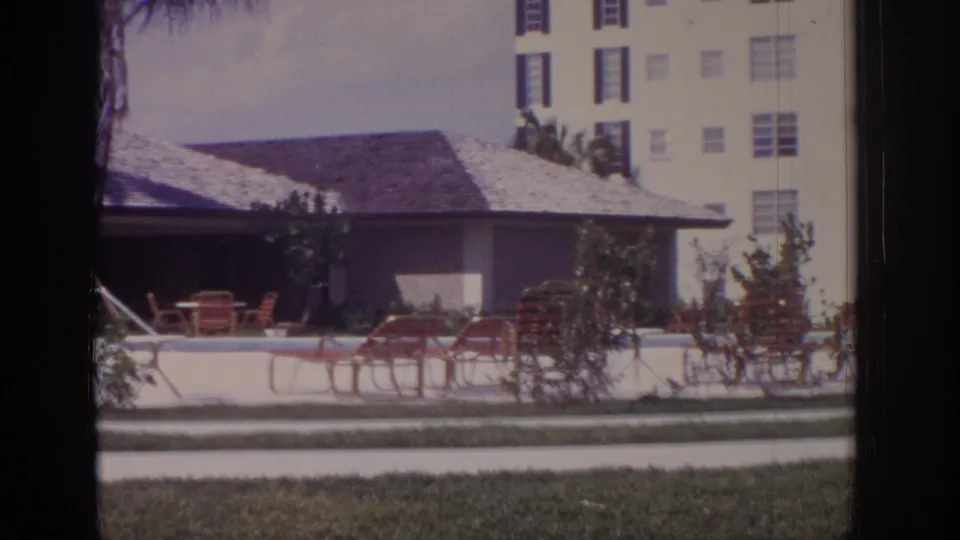 1973 Naples, Florida frame with a low tiled-roof resort building, poolside lounge chairs, landscaped shrubs, lawn, walkway, and a taller hotel block behind it.