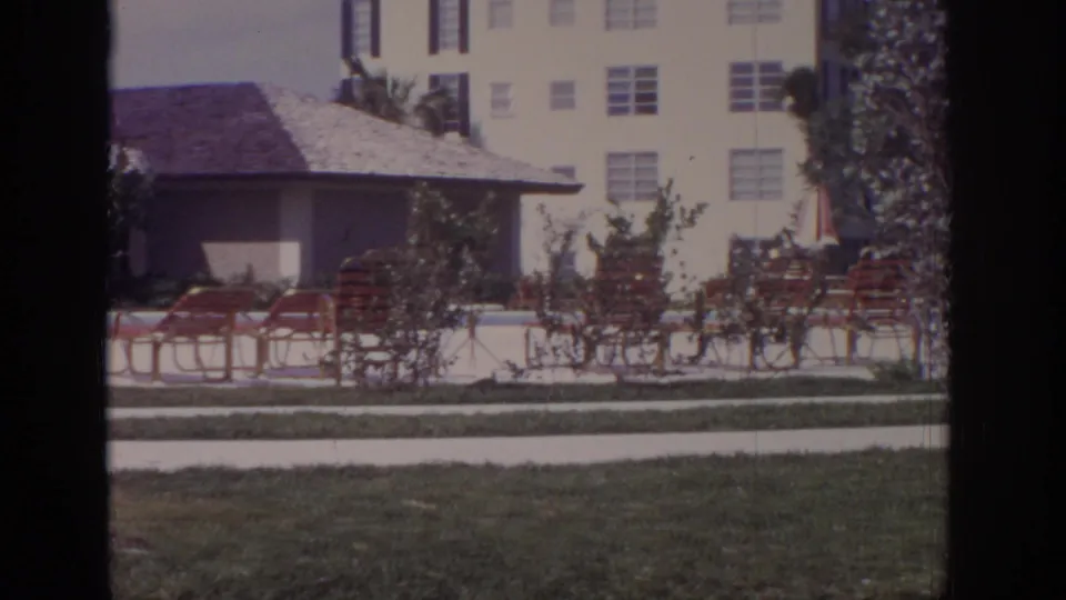 End frame of the 1973 Naples, Florida poolside pan showing red lounge chairs, a striped umbrella, shrubs, palms, a low resort building, and a multi-story hotel facade.