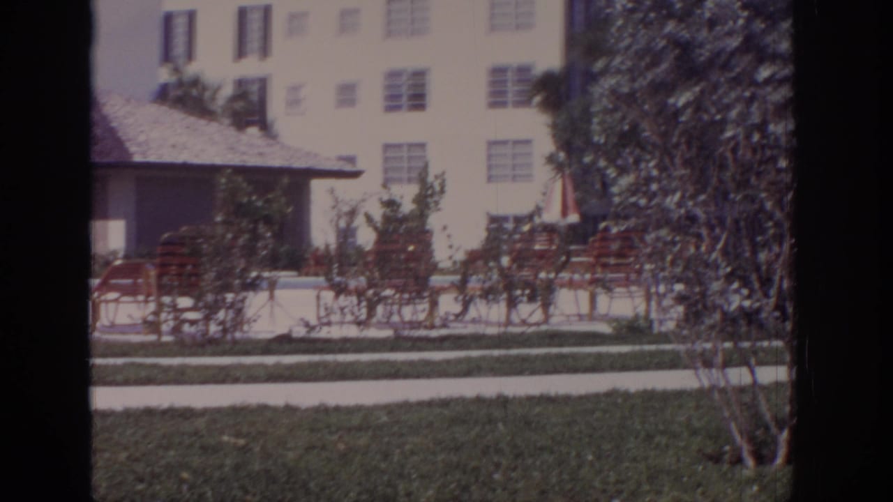 1973 Naples, Florida home-movie frame showing a poolside residential area with chairs, grass, shrubs, cabana roof, and apartment buildings.
