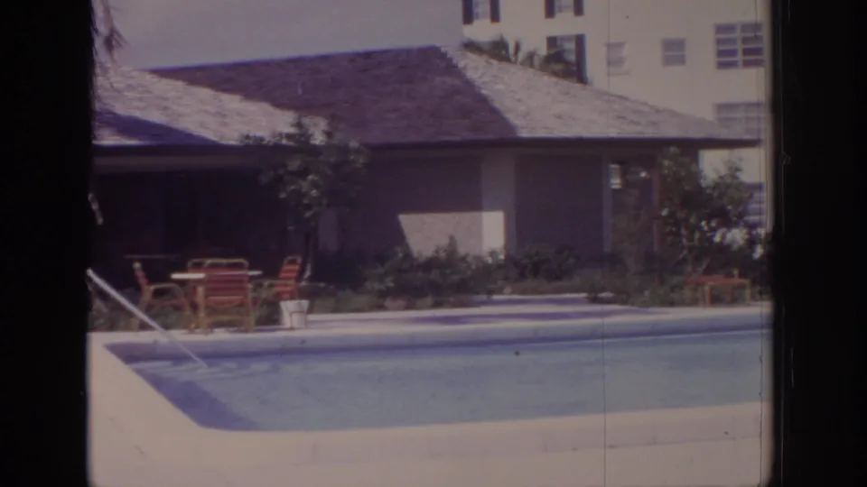 1973 Naples, Florida poolside view with blue water, a cabana-like structure, lounge furniture, planted landscaping, and apartment buildings.