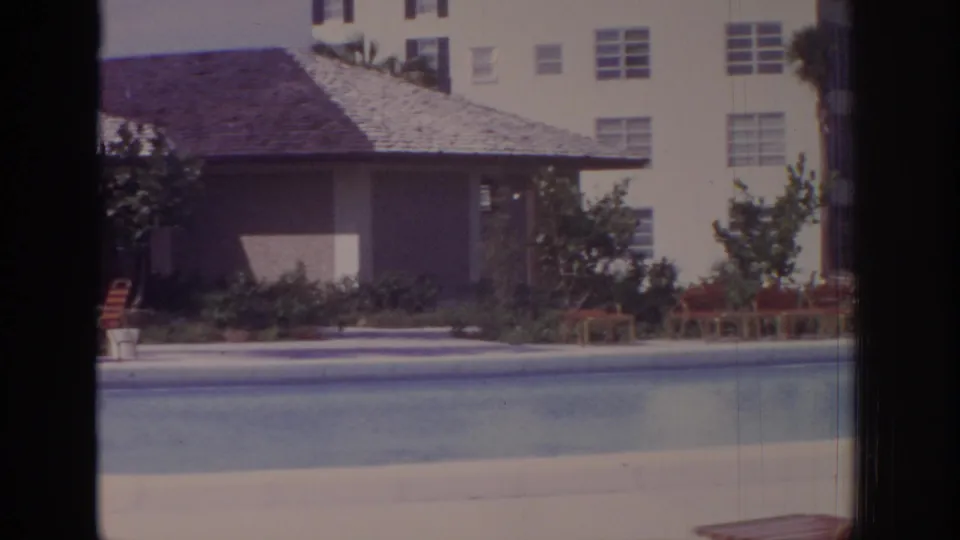 1973 Naples, Florida frame showing the residential pool foreground, shaded cabana roof, lounge furniture, shrubs, and a pale apartment building.